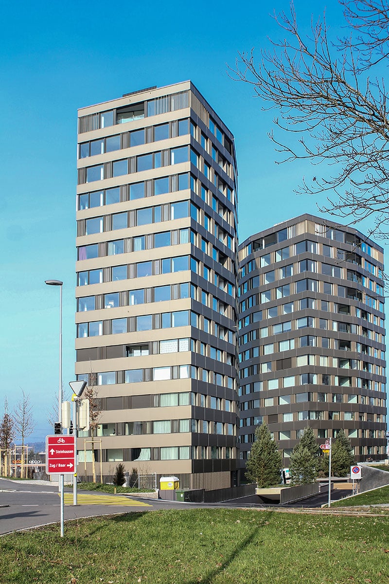 Multi-story apartment building with a modern, angular design featuring large windows and a mix of light and dark tones. There is a parking area visible in the foreground.