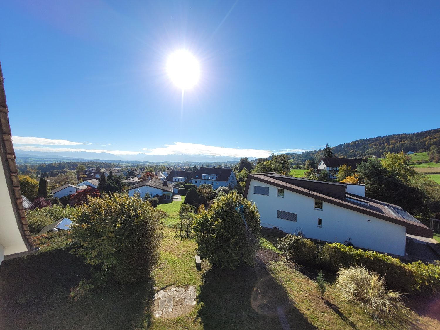 Multiple houses with green lawns, trees, hills and mountains in the background, bright sun shining