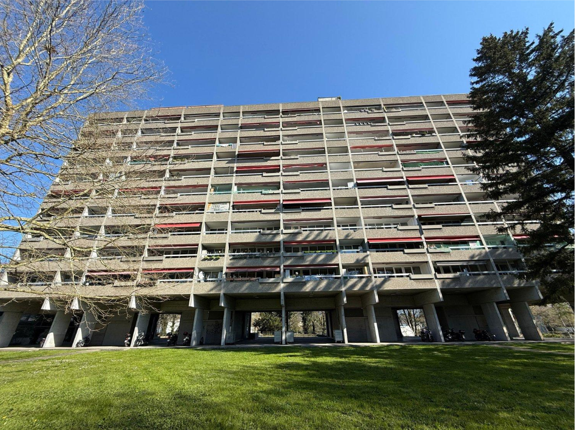 A large multi-story concrete building with a complex facade featuring various window sizes and patterns. The building is surrounded by a grassy lawn and trees.
