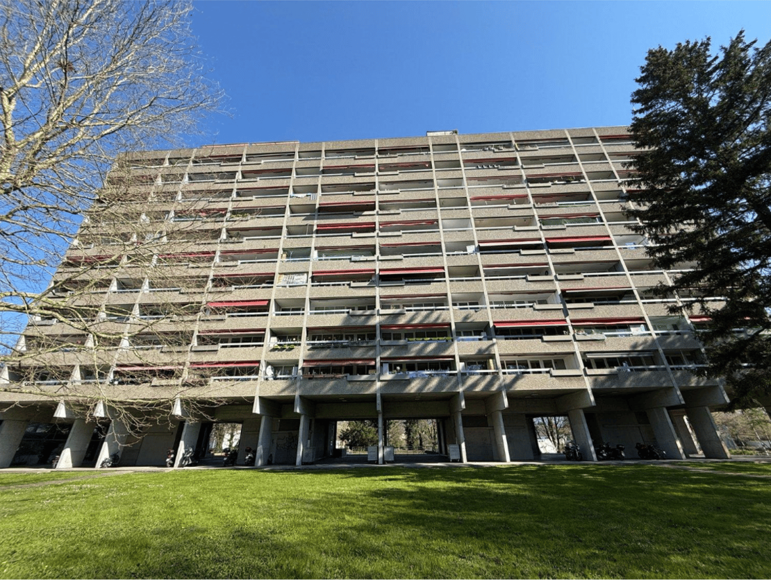 A large multi-story concrete building with a complex facade featuring rows of windows and balconies. The building is surrounded by a grassy lawn and trees.