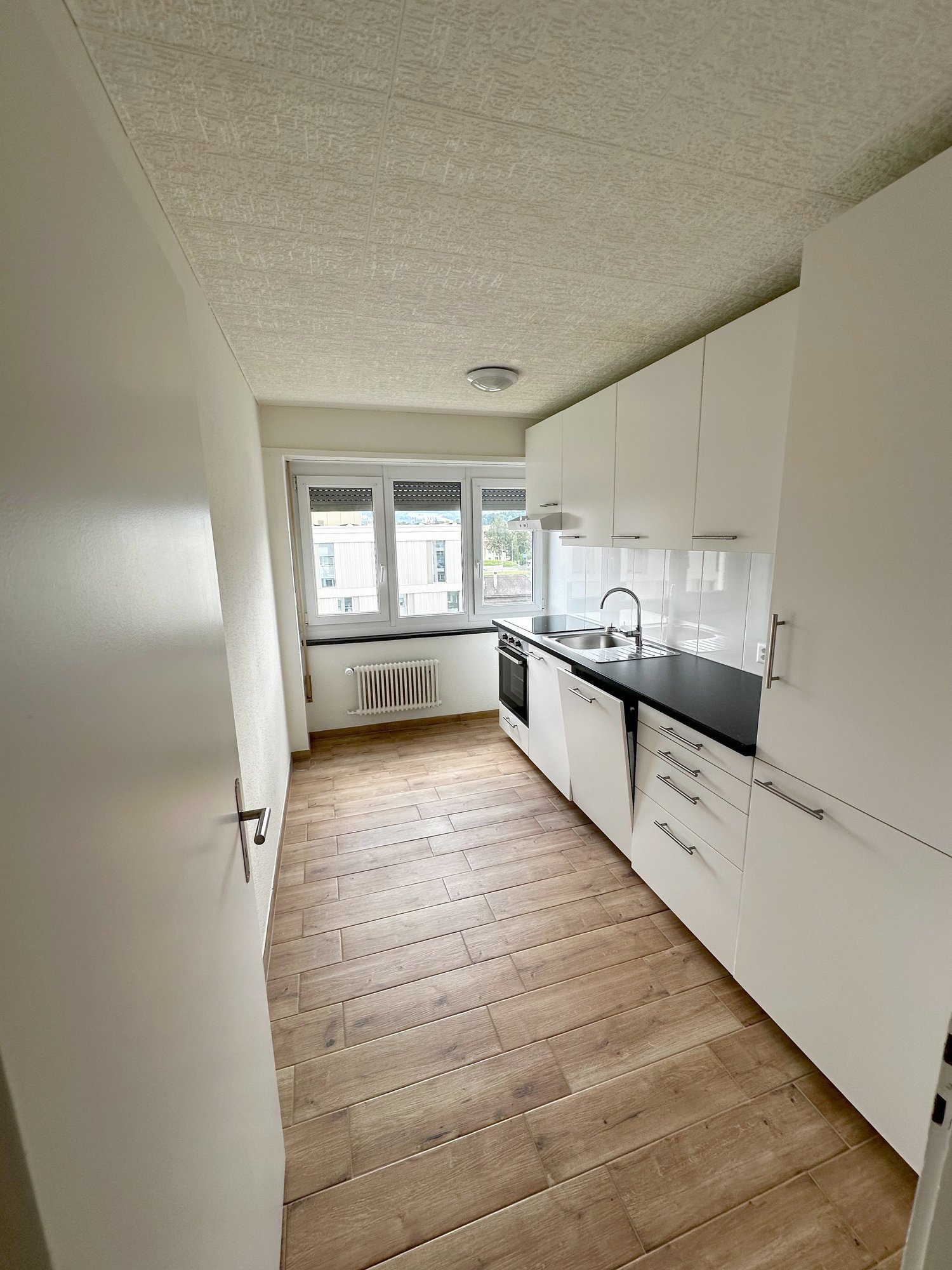 empty kitchen with white cabinets, wooden floor, black countertop, sink with faucet, oven, window