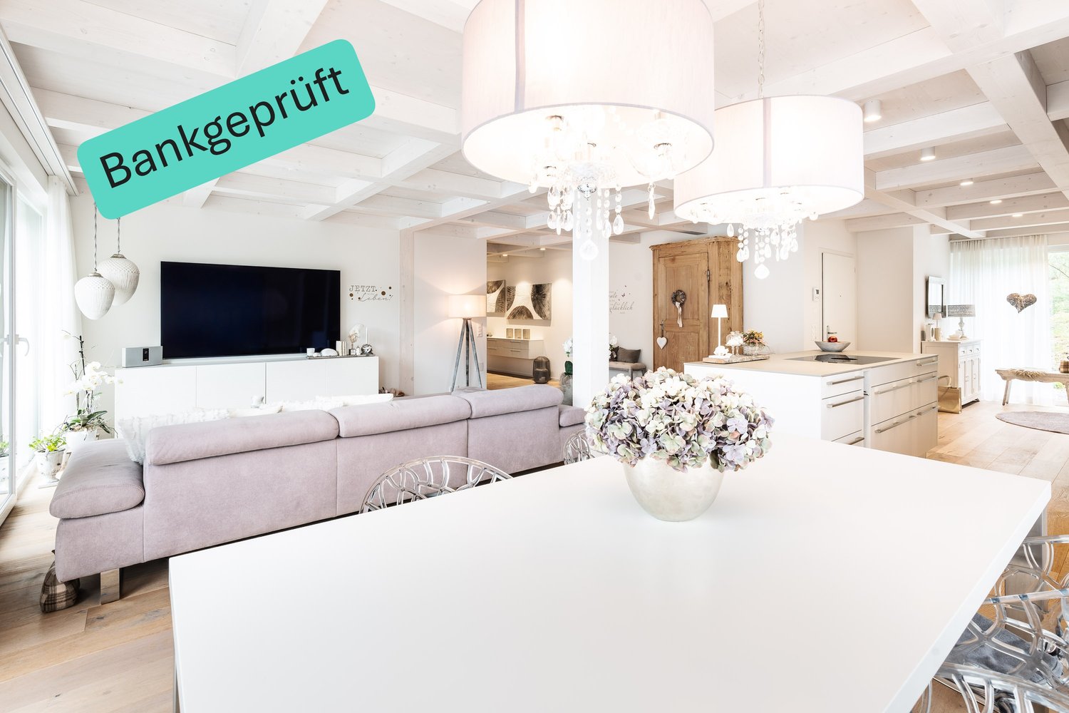 living room with white walls, wooden floor, ceiling with wooden beams, chandeliers, couch, dining table with flower vase, flat-screen TV, white kitchen with island, white cabinets, glass windows