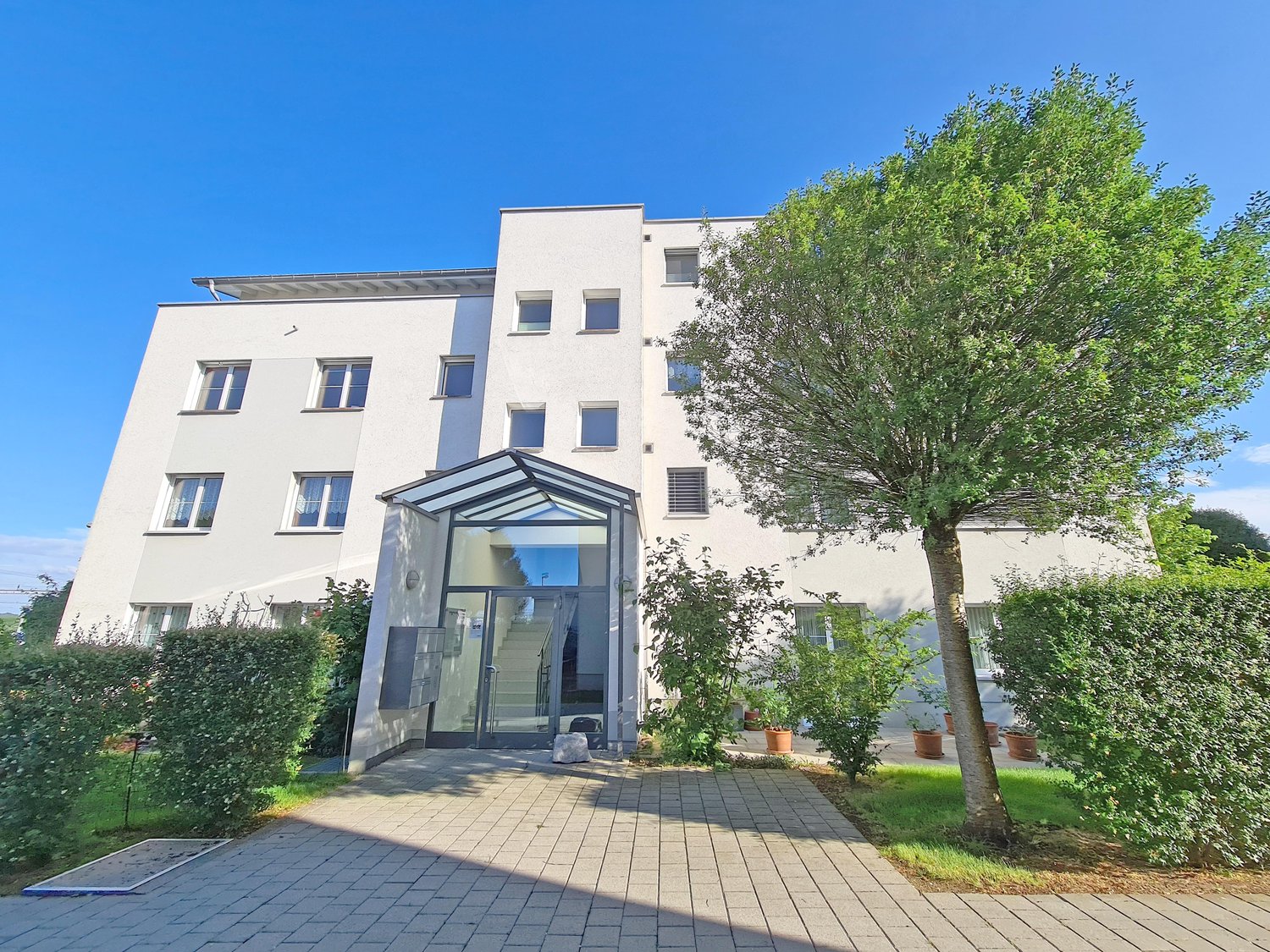 Multi-storey residential building, white facade, glass door, green trees, bushes, brick walkway, windows, stairs