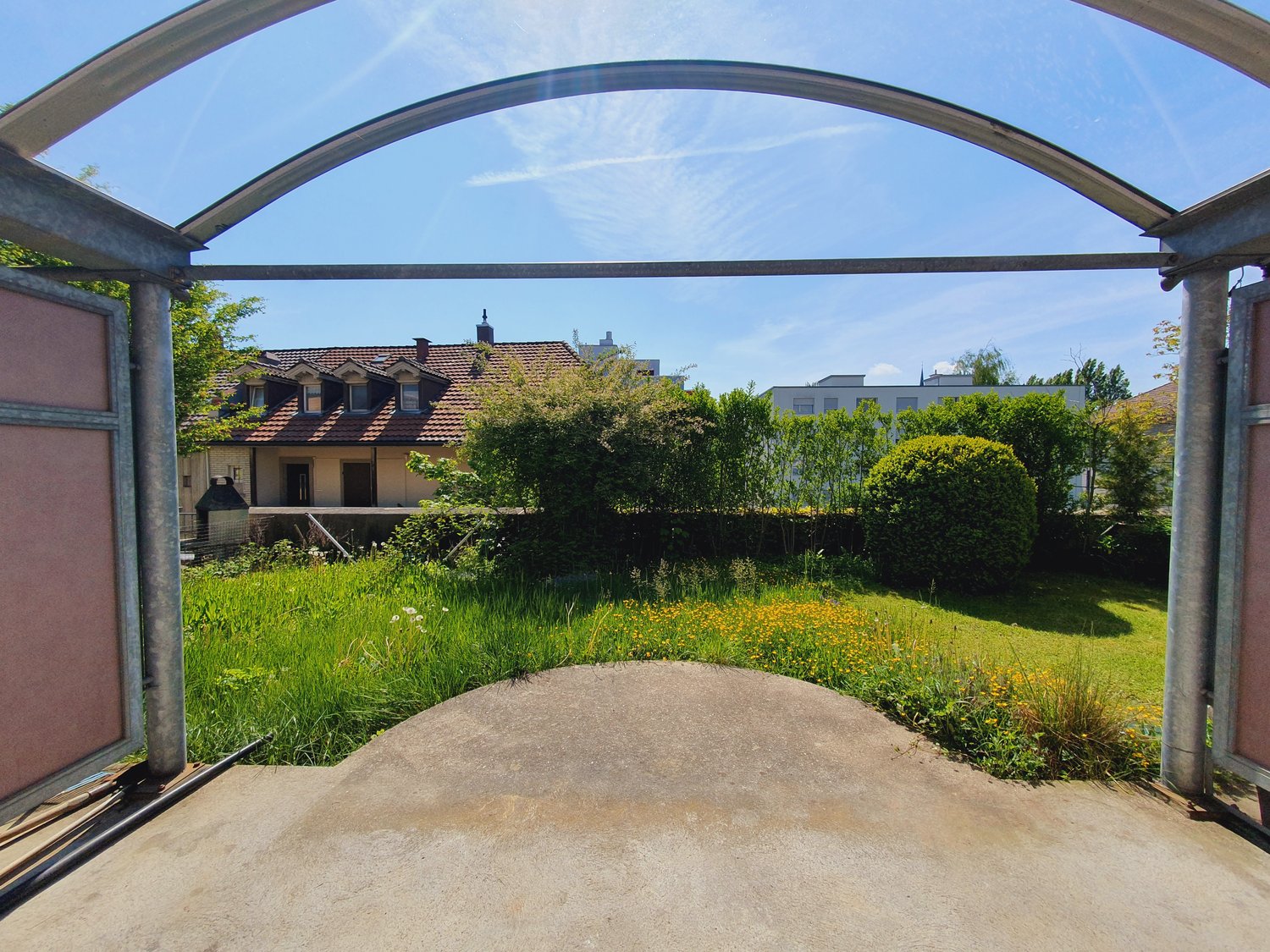 Covered parking area leading to the house's backyard, surrounded by grass and shrubs.