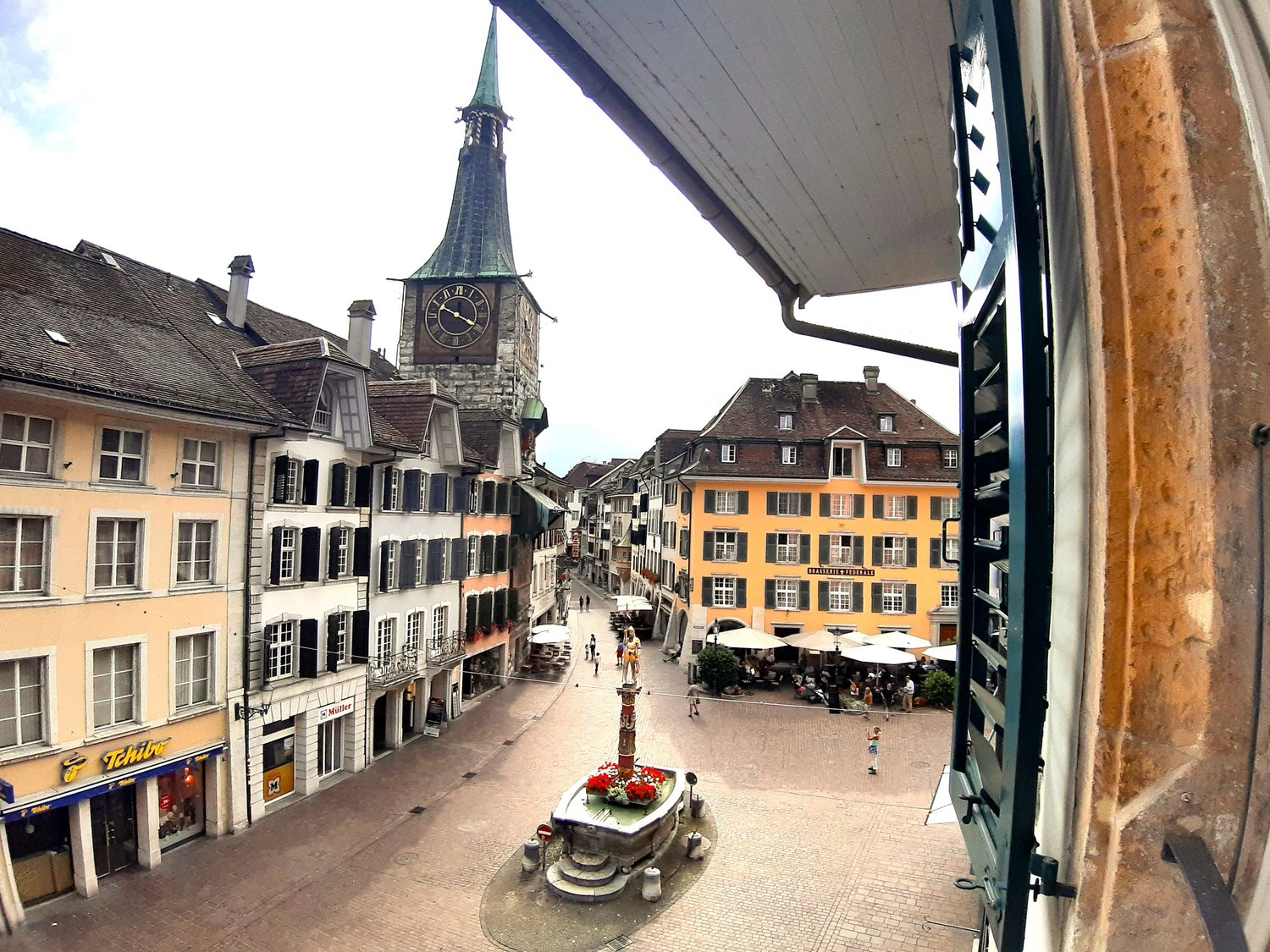 Church clock tower, street with shops, square with fountain, buildings, cobblestone streets, pedestrians