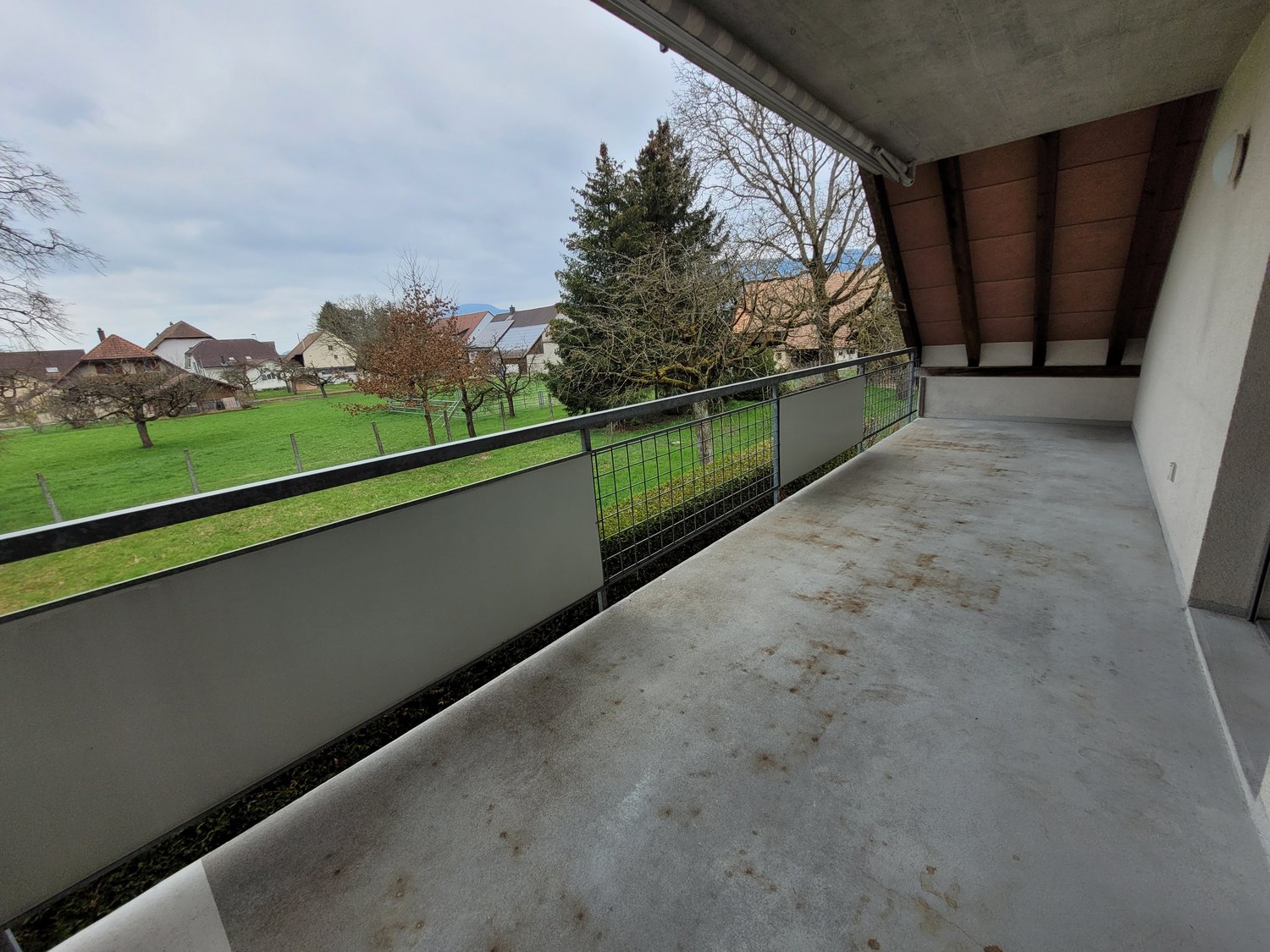 large balcony with metal railing, overlooking a grassy field and houses