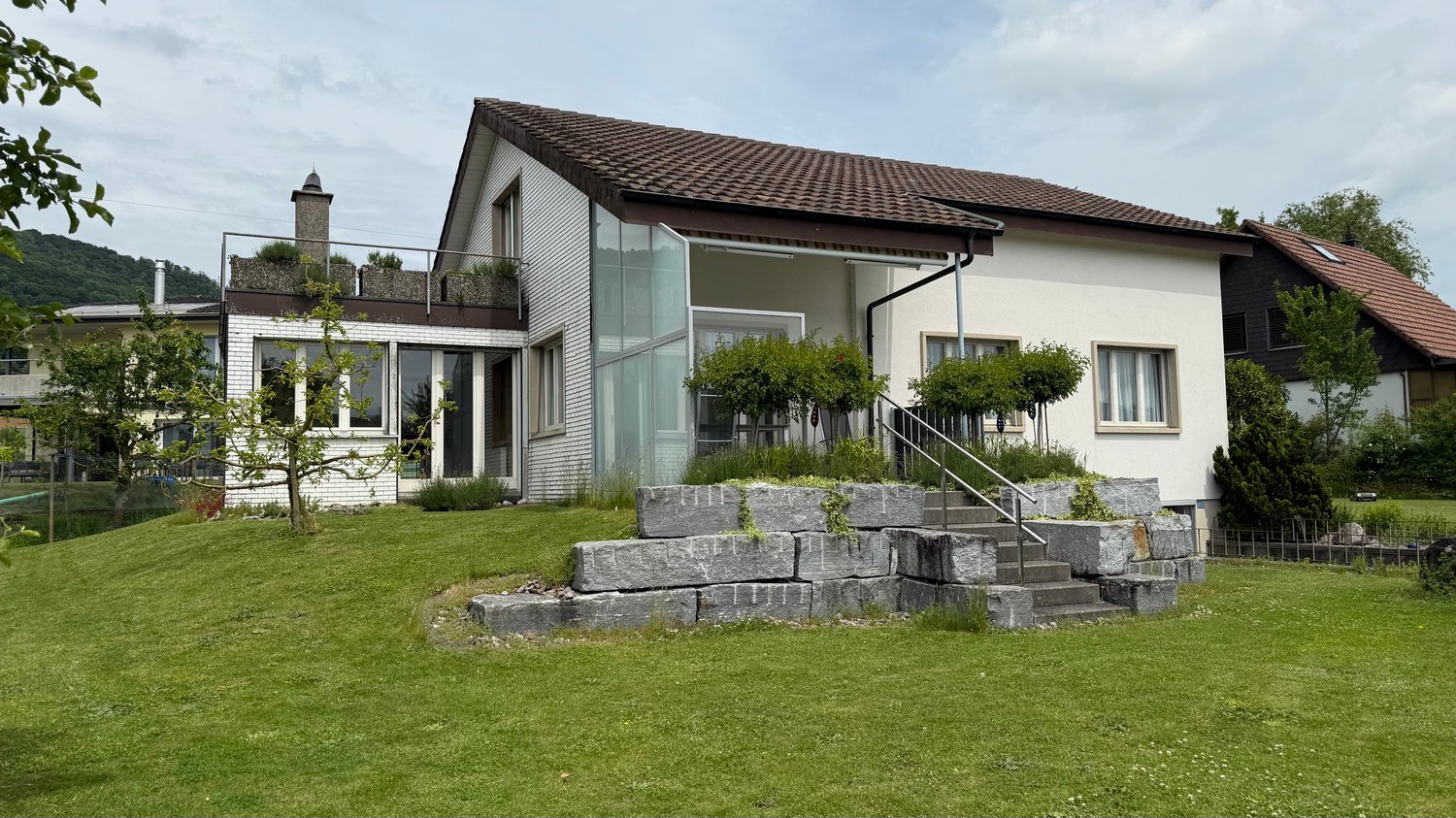Raised ground floor on hillside, brown roof, large windows, green lawn, view of mountain