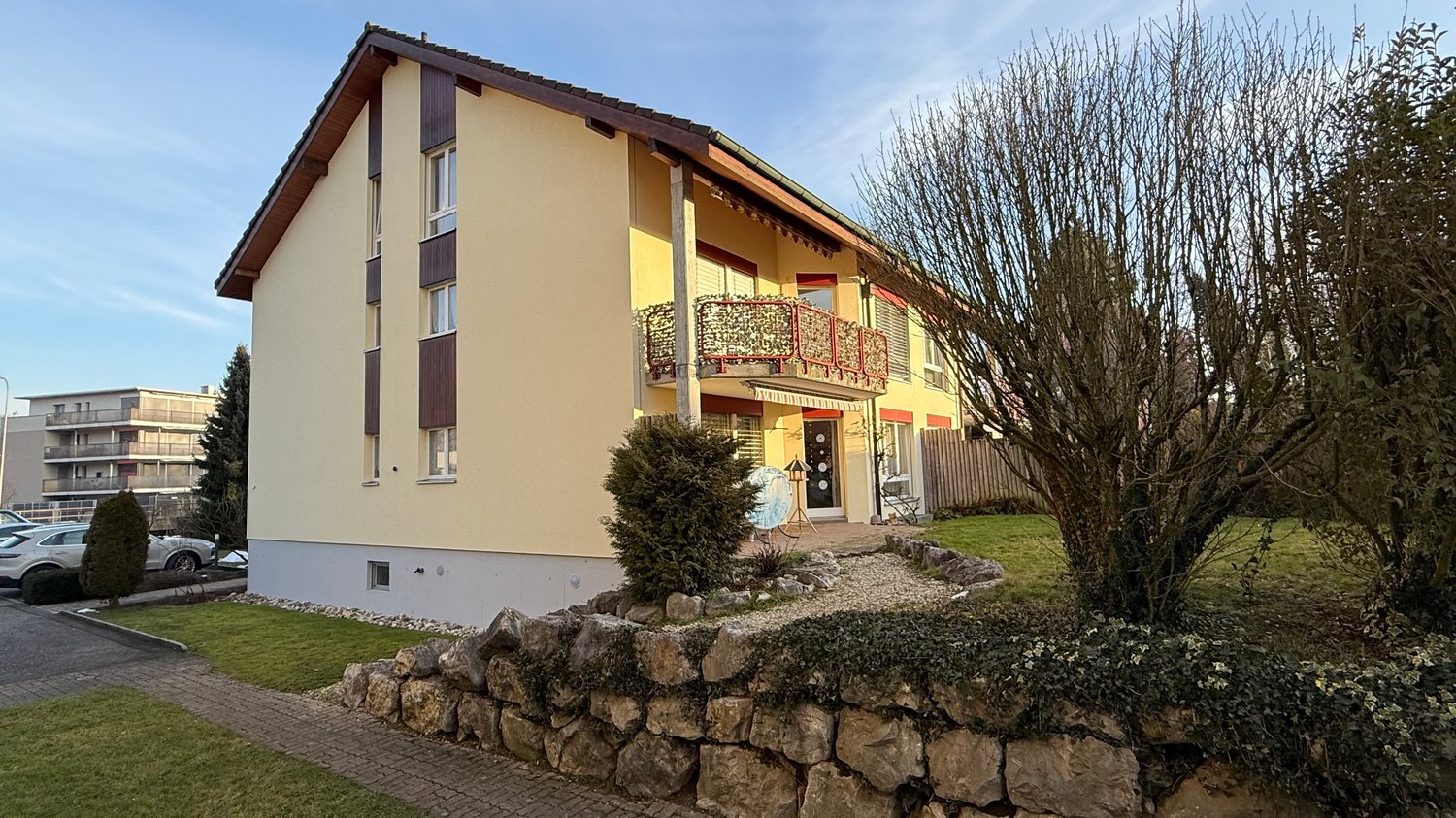 Two-story yellow house with a balcony, brown roof, ground-floor terrace, and a stone wall in the front garden.