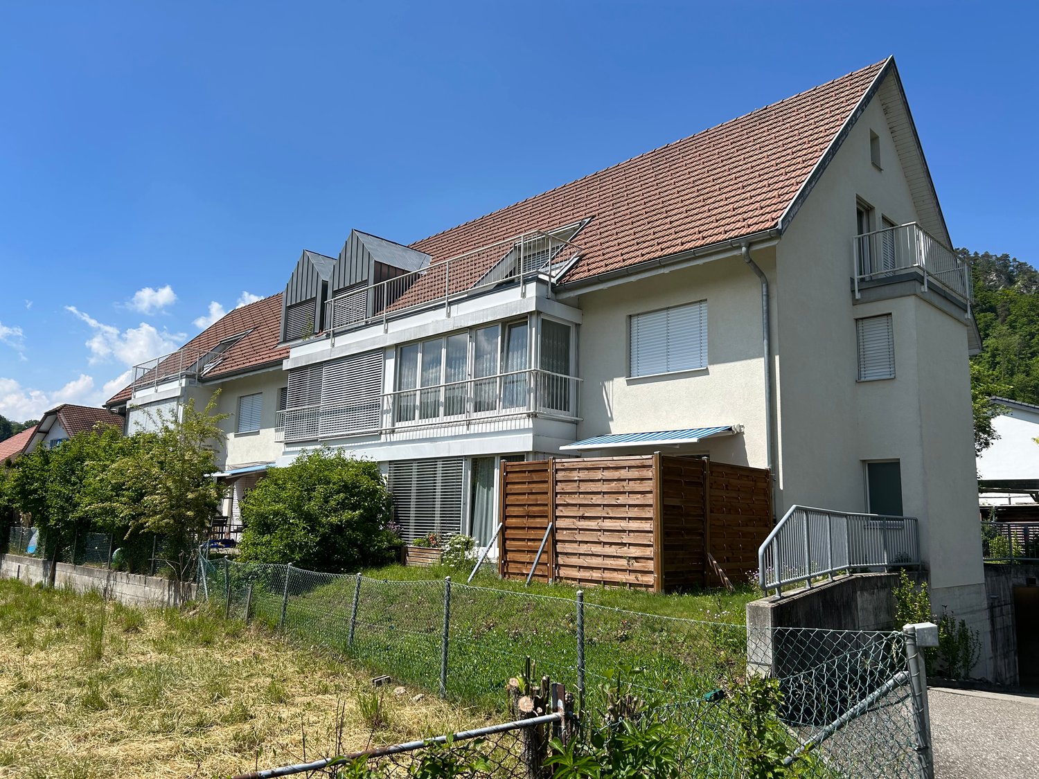 3 story house, white exterior, red roof, multiple balconies, solar panels, fenced yard, wooden fence, trees, plants