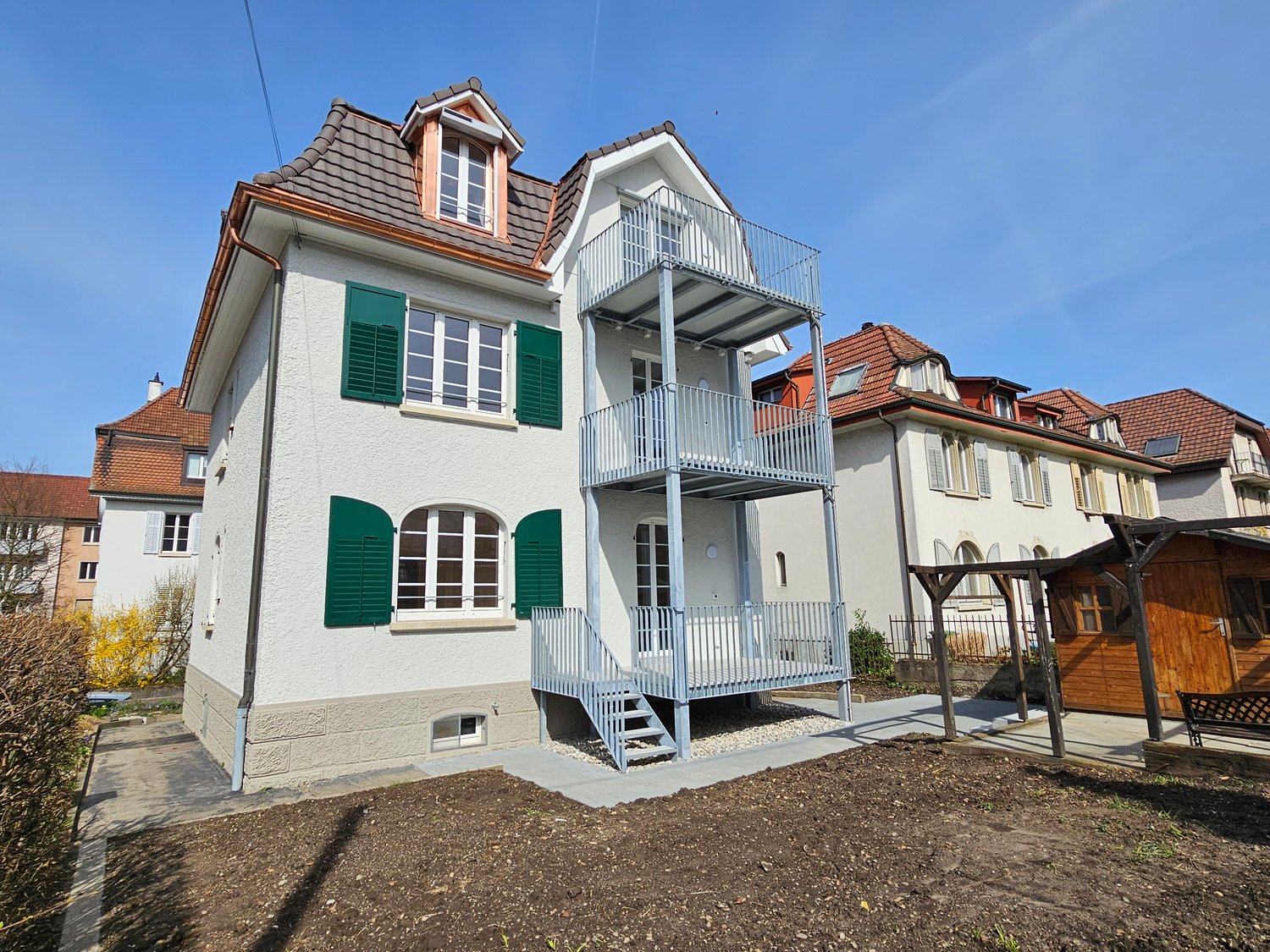Two-story house, white walls with green shutters, brown roof, staircase leading to balcony