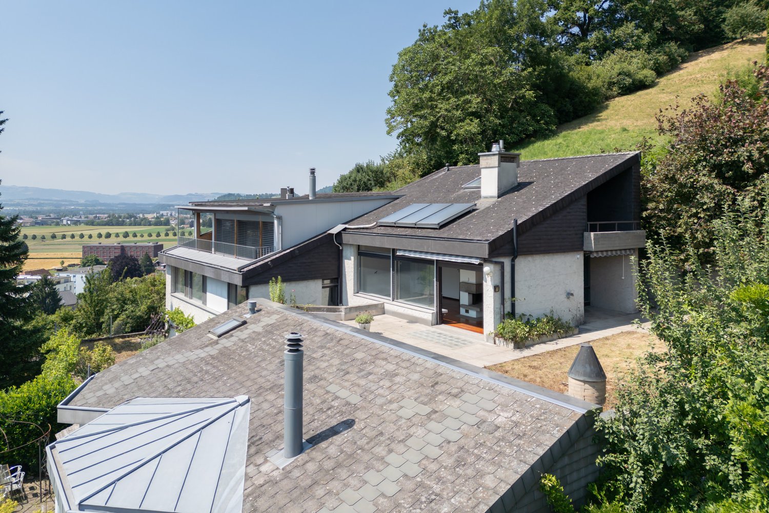 Two-story modern house with a slanted roof, large windows, and a balcony overlooking a scenic landscape with trees and hills in the background. The house has a paved driveway or patio in the front.