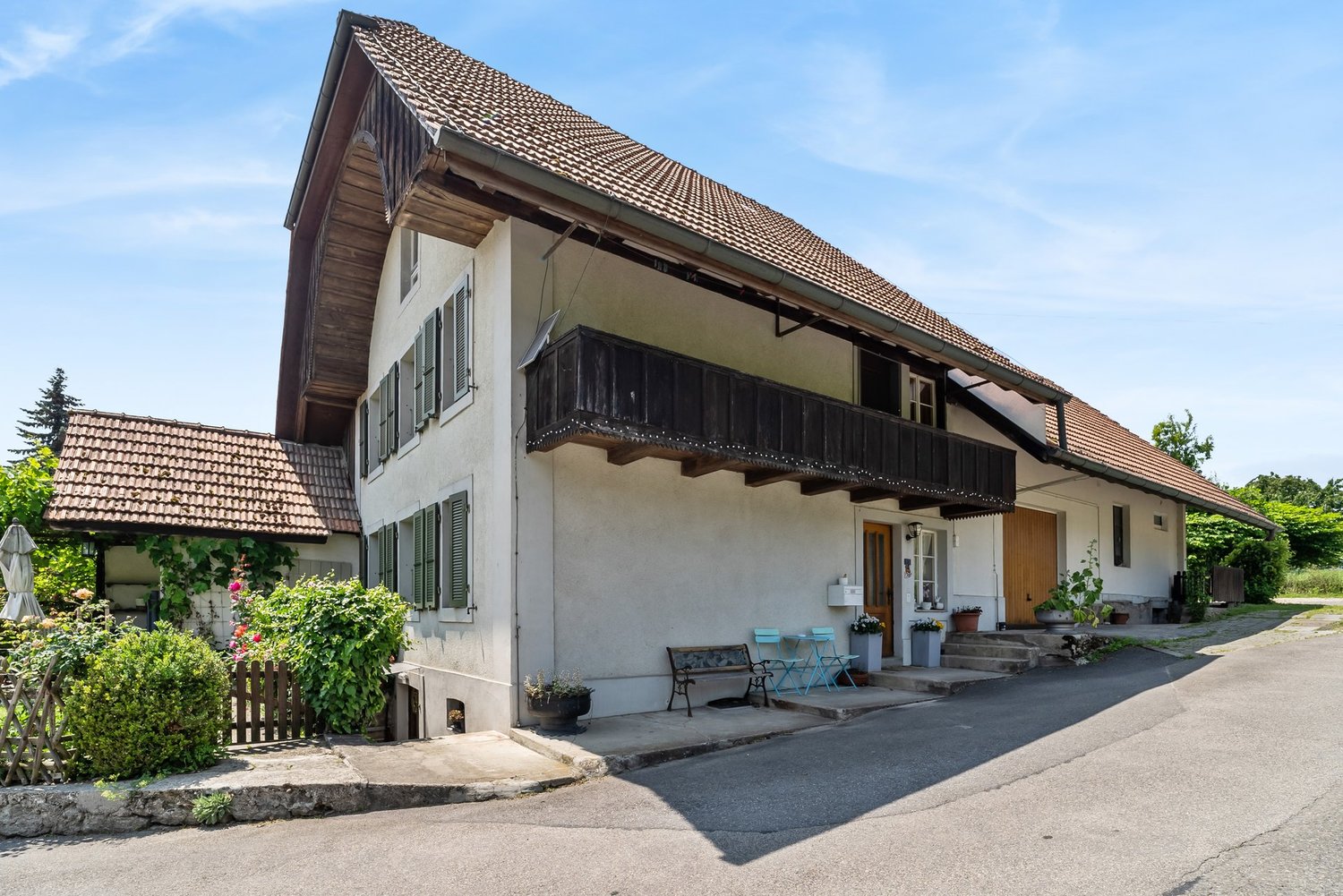 2-story house with a wooden balcony, stone walls, and a tiled roof. The house has a paved driveway and is surrounded by greenery.