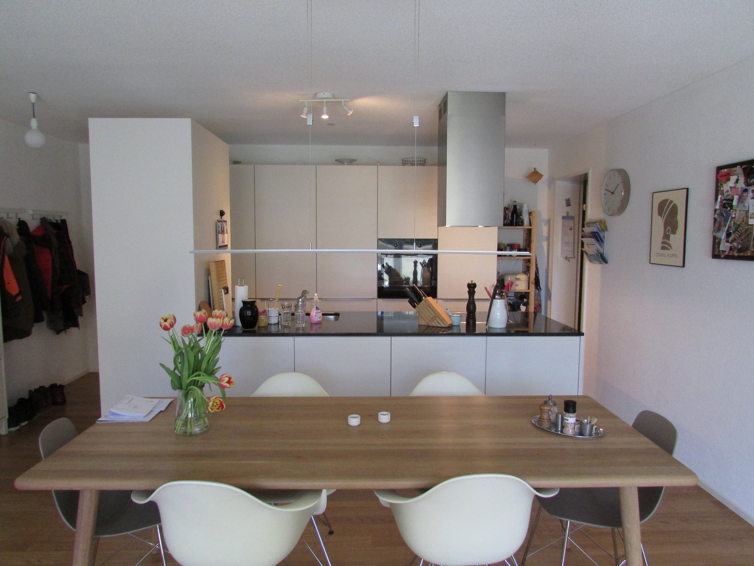 The image shows an open-concept kitchen and dining area. The kitchen features white cabinets, a stainless steel appliance, and a dishwasher. The dining area has a wooden table with white chairs. There are various decor items such as tulips in a vase, artw