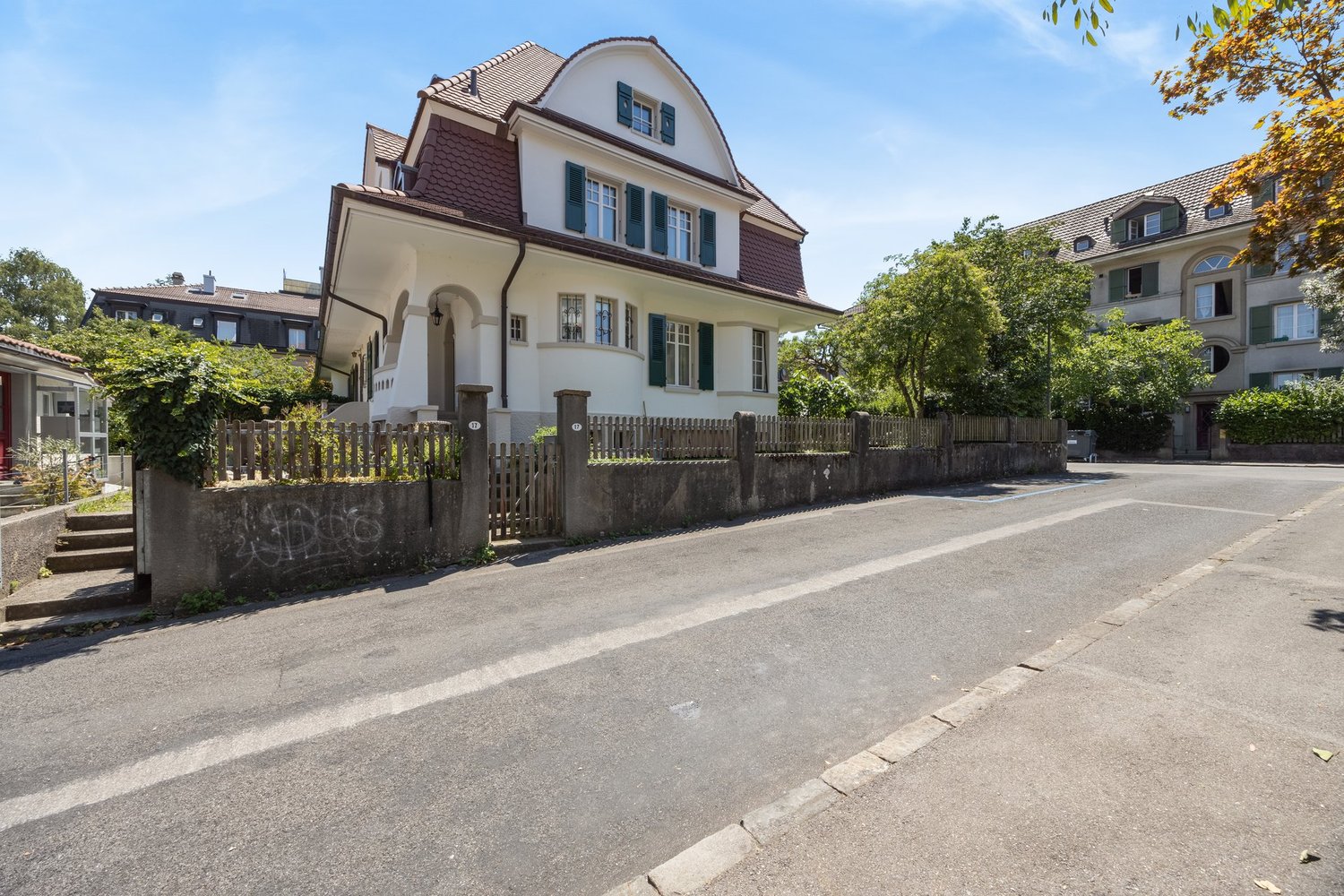 A two-story house with a brown roof, white walls, and green shutters. It features a spacious garden with a wooden fence and trees. In front of the house, there is a clear street with no cars, marked with a white line. The house is surrounded by other buil