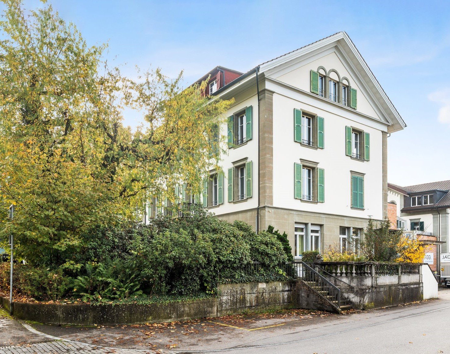 Three story building, white facade, green shutters, balcony, trees and bushes in front