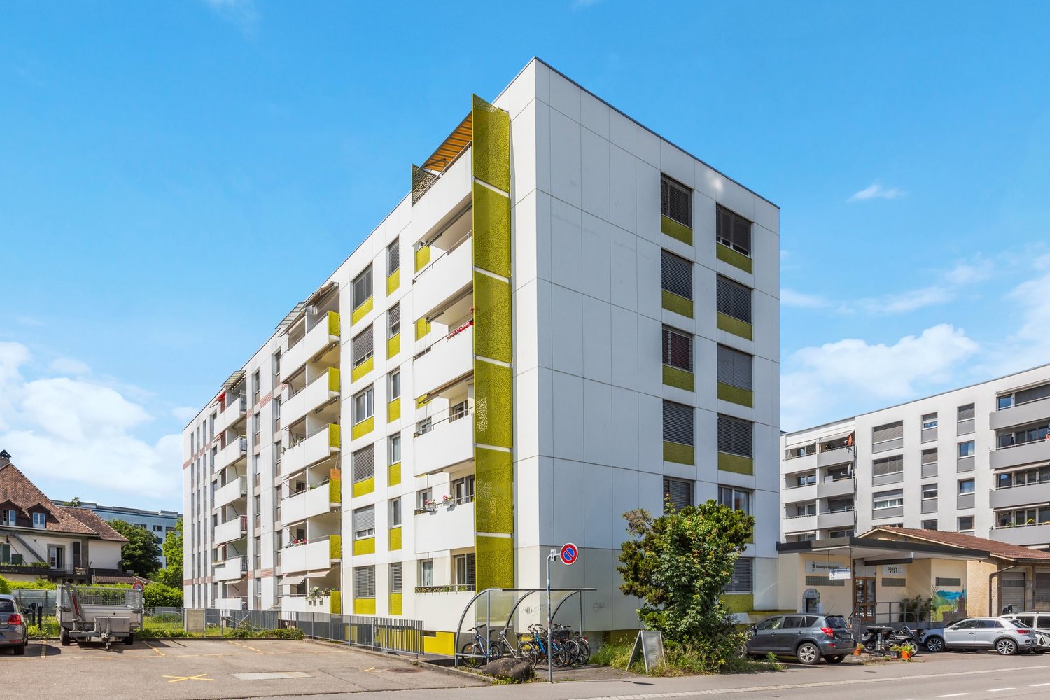 Multi-storey apartment building with yellow accents, several windows, balconies, and a parking lot with bicycles and cars.