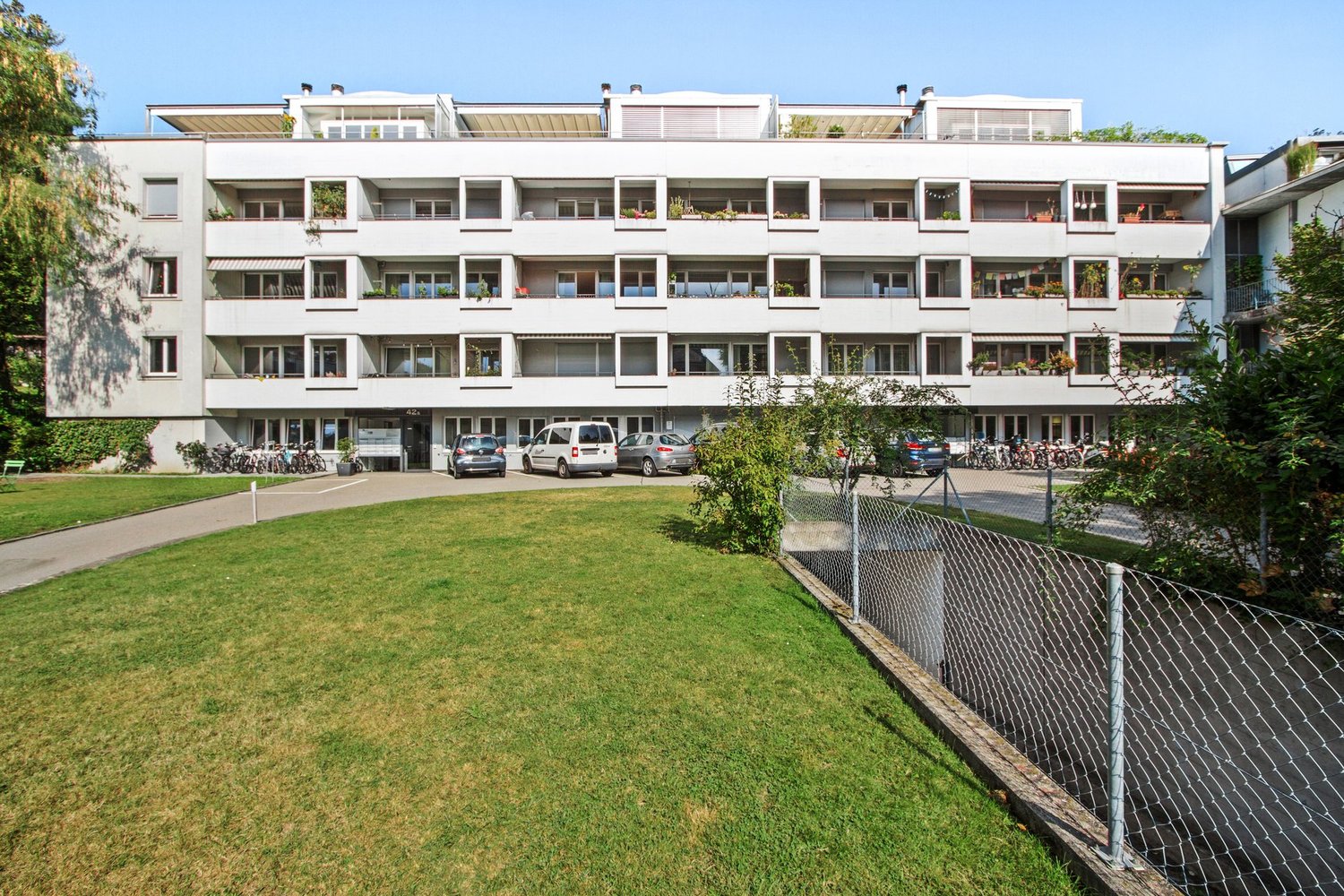 multistory building, white exterior, multiple balconies, several cars parked in front, fenced lawn
