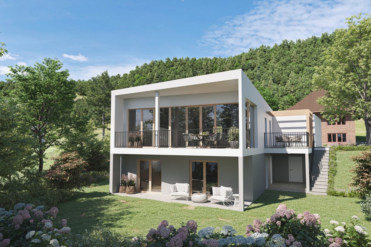 Two-story modern house with a flat roof, large windows, and a balcony on the upper level. The house is surrounded by a well-manicured lawn and lush greenery, including trees and flowering plants. There appears to be a garage attached to the side of the ho