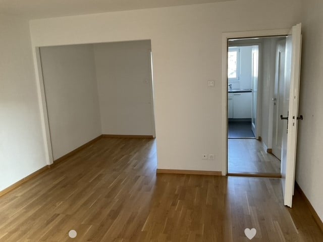Empty room with wooden floors, white walls, and two open doors leading to a kitchen.