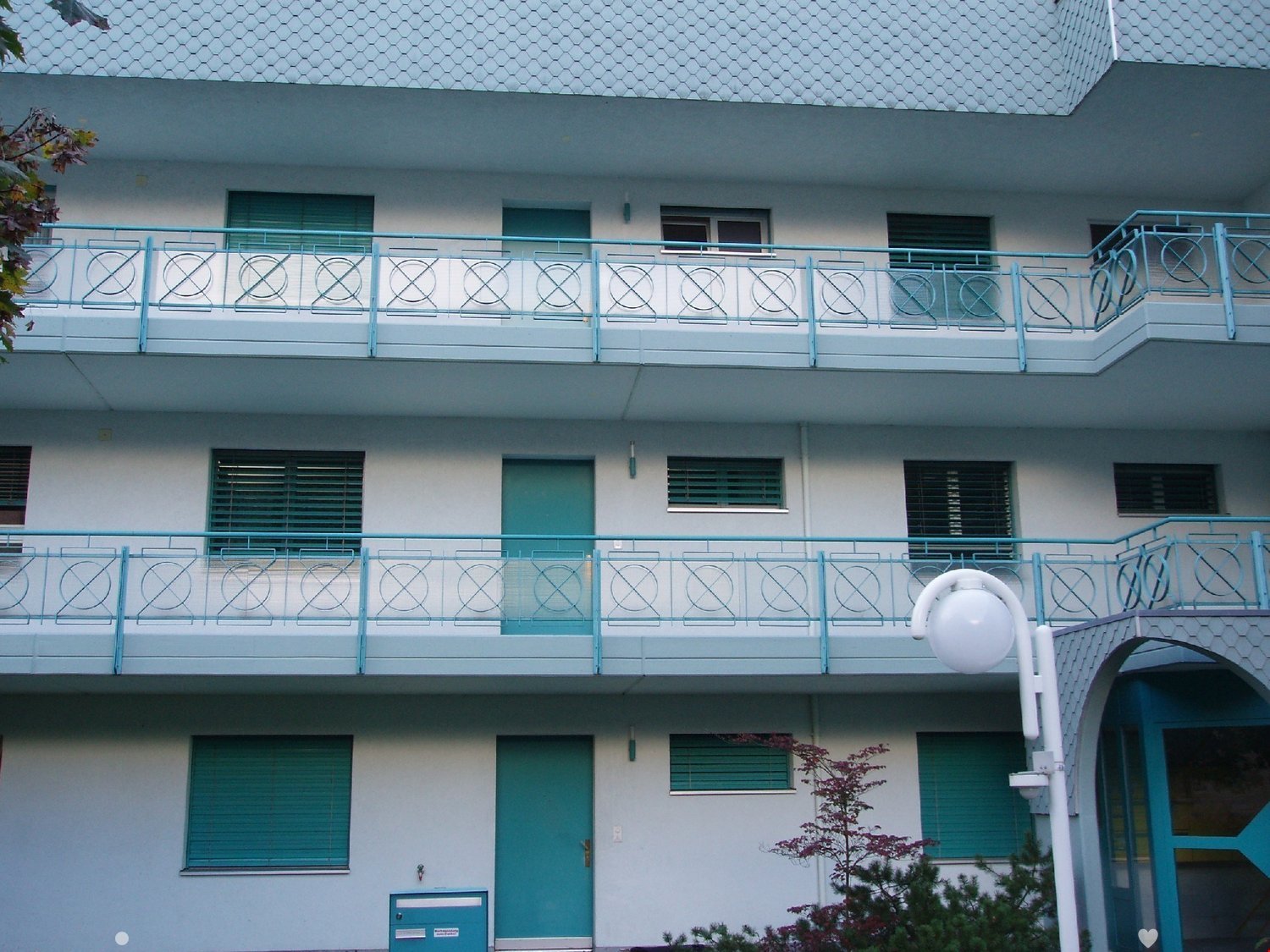 Three story apartment building, white walls, blue balconies, blue shutters, brick roof