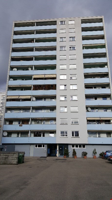 Modern apartment building with multiple floors, balconies, windows, satellite dishes, plants in front.