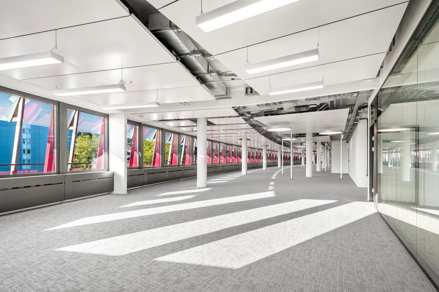 Large open office space with high ceilings, exposed ductwork, and floor-to-ceiling windows providing natural light. The space has a modern, minimalist design with grey carpeting and white walls. There are multiple columns throughout the space.