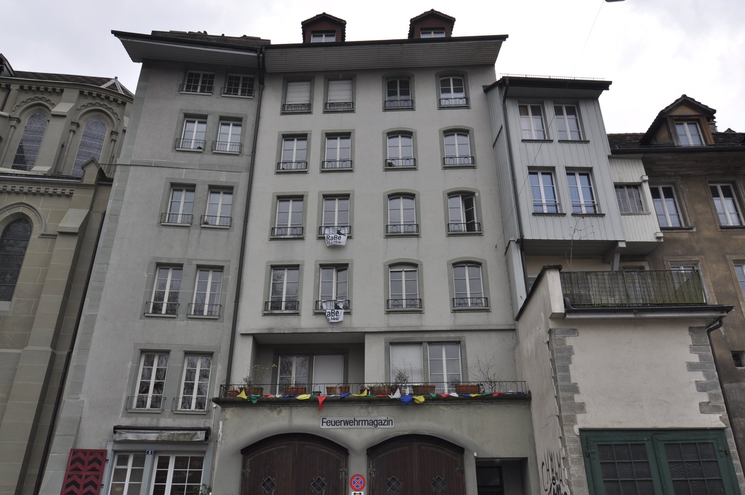 Multi-story building with a mix of architectural styles, including arched windows and balconies. The building has a sign that says 'Feuerwehrmagazin', indicating it may be a fire department or related facility. The building has a gray exterior and appears