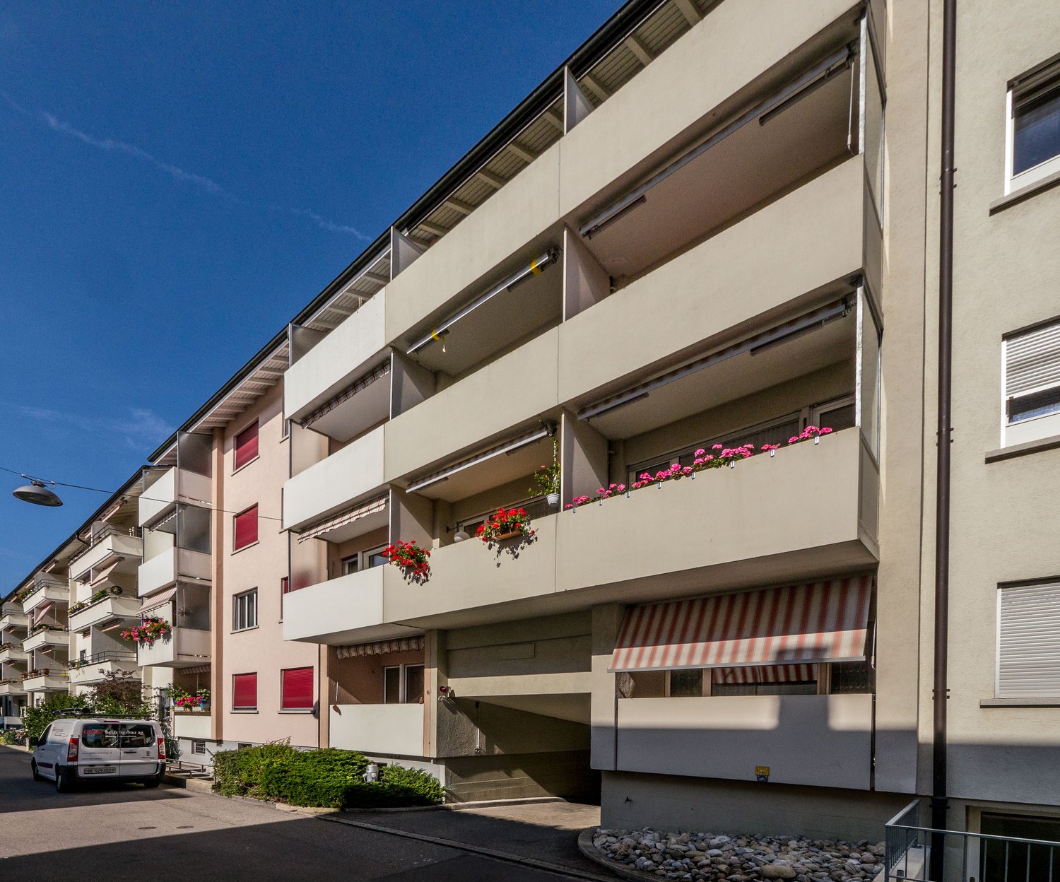 Multi-story building with balconies, red trim around windows, street view, white vans parked in the street