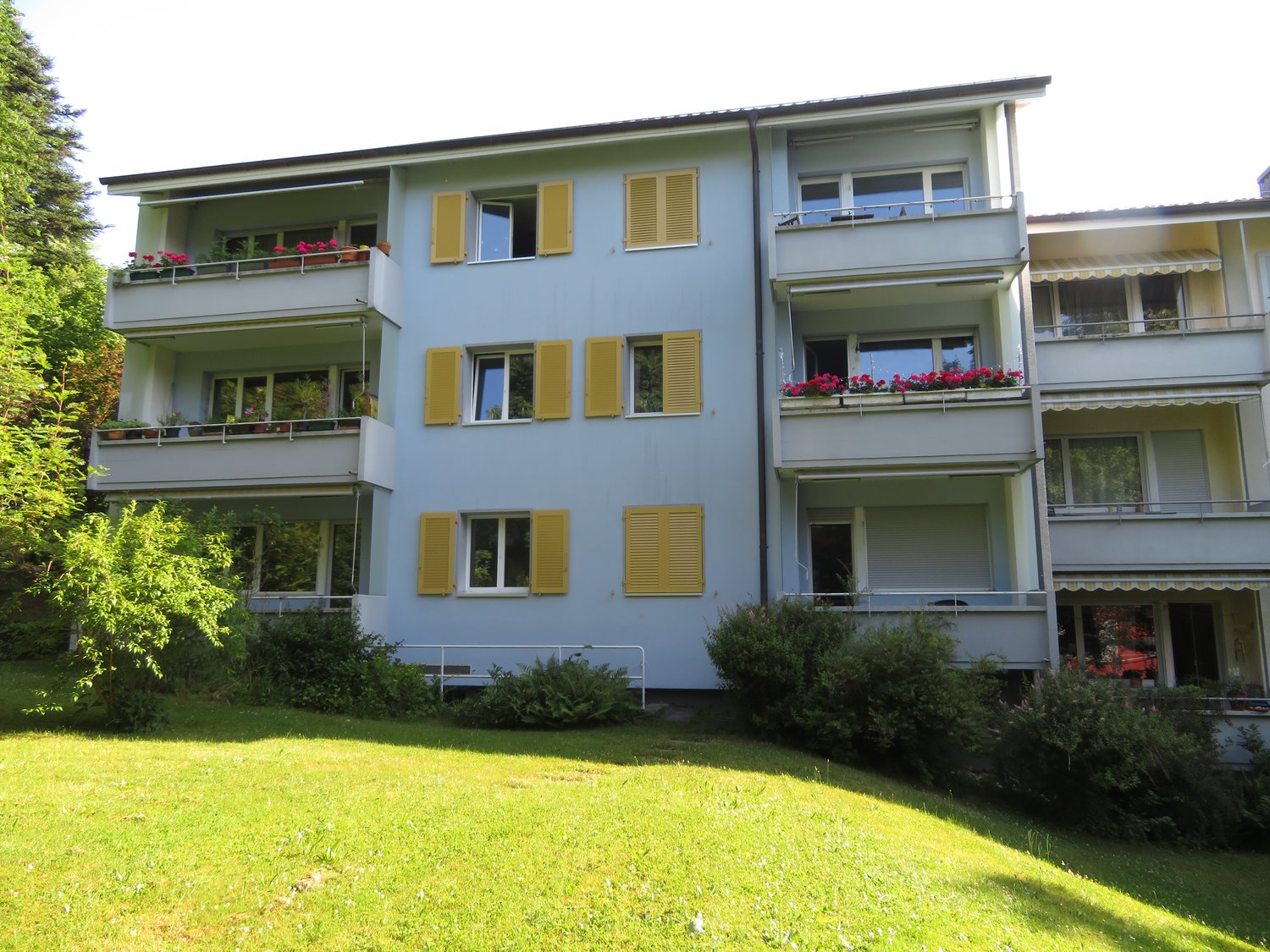A four-story building with yellow shutters, balconies with potted plants and flowers, and a small green lawn in front