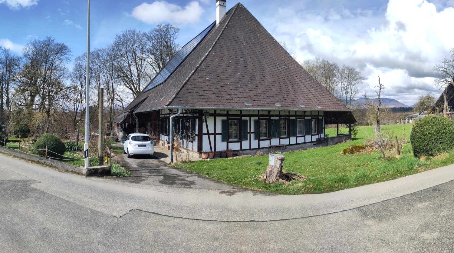 A traditional German-style half-timbered house with a large pyramidal roof, surrounded by trees and a grassy area. There is a parked car in the driveway in front of the building.