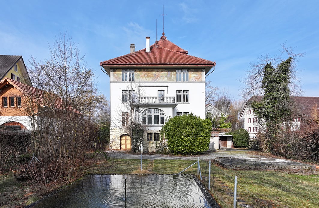 3-story historic building with red tiled roof, stone facade, large windows, balcony, surrounded by trees and a small pond in the foreground