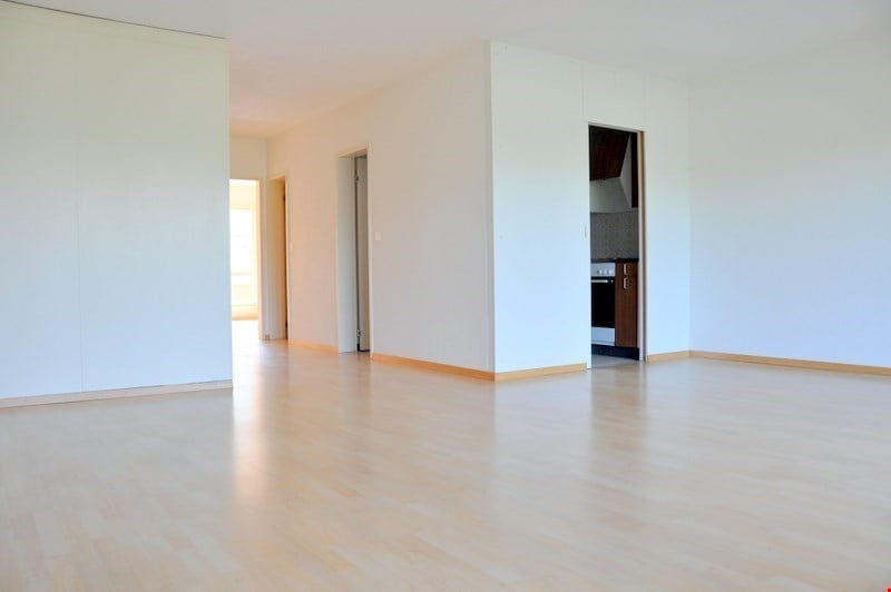 Empty room with wooden floor, white walls, kitchen with stove and wooden cabinets.