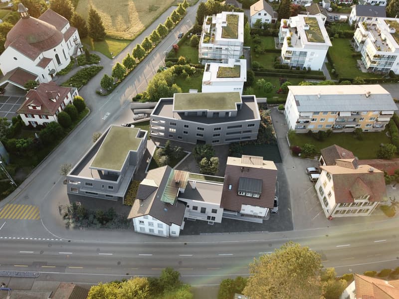 The image shows an aerial view of a residential area with a mix of multi-story apartment buildings, townhouses, and single-family homes. The buildings have a modern, contemporary design with flat roofs and large windows. There are paved roads and parking