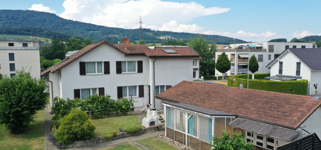 Multi-story apartment building with white exterior walls, red tile roofs, and balconies. The building is surrounded by trees and greenery, with other residential buildings visible in the background.