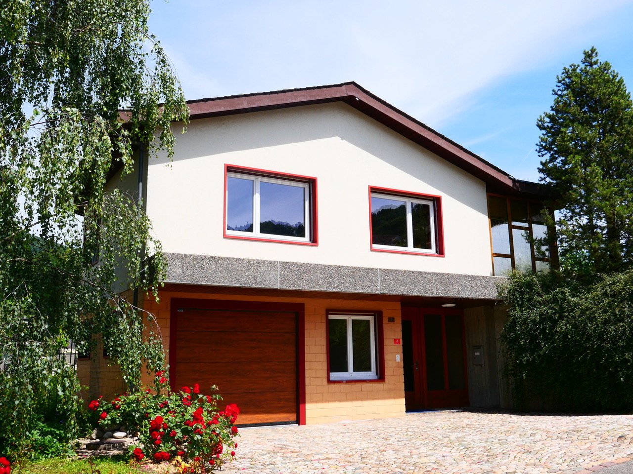 2-story house with white exterior walls, red-framed windows, and a wooden garage door. The house has a balcony or terrace on the upper level. The front yard features a paved driveway and red flowers in the landscaping.