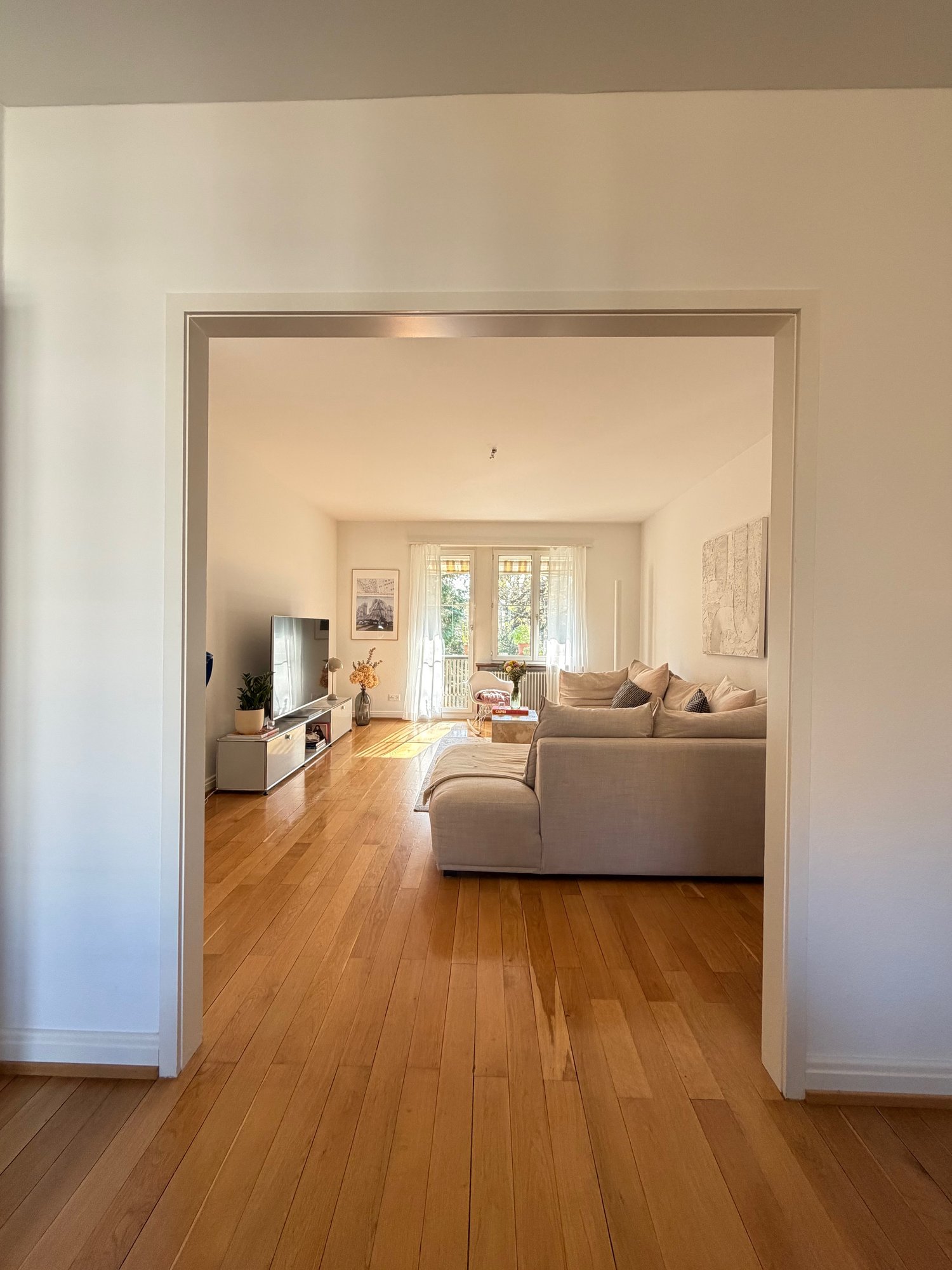 Spacious open plan living room with hardwood floors, white walls, a balcony with an elegant glass door, a comfortable beige sofa, a TV on a cabinet, and a glass center table with flowers. There is also a potted plant, a desk with a lamp, and a framed pict