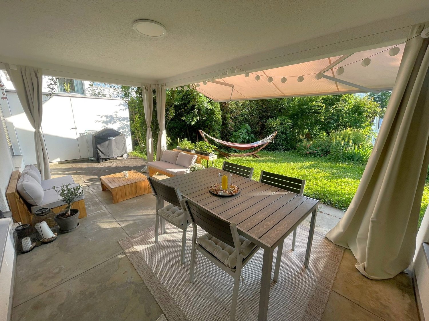 Covered patio with wooden dining table and chairs, sofa, hammock, and string lights. The patio overlooks a lush, green garden with trees and plants.