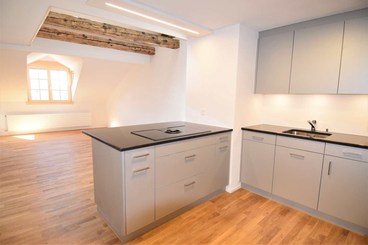 Empty kitchen with central island and black counter top, white cabinets, light wood floor, sink and faucet