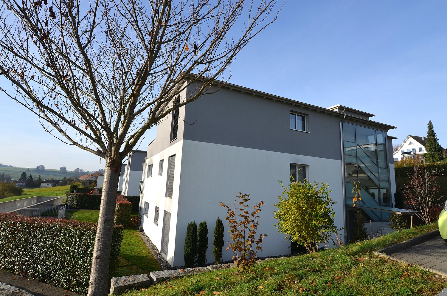 Modern two-story building with a flat roof, white exterior walls and gray trim, glass windows and doors, a small glass-covered entrance, surrounded by a garden with hedges and plants.