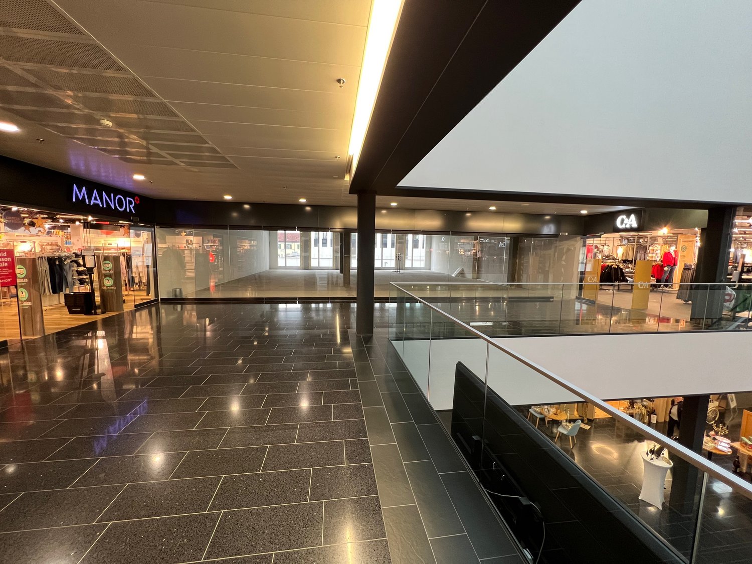large shopping mall, empty, tiled floor, high ceiling, shops, sign for MANOR, light and glass railings