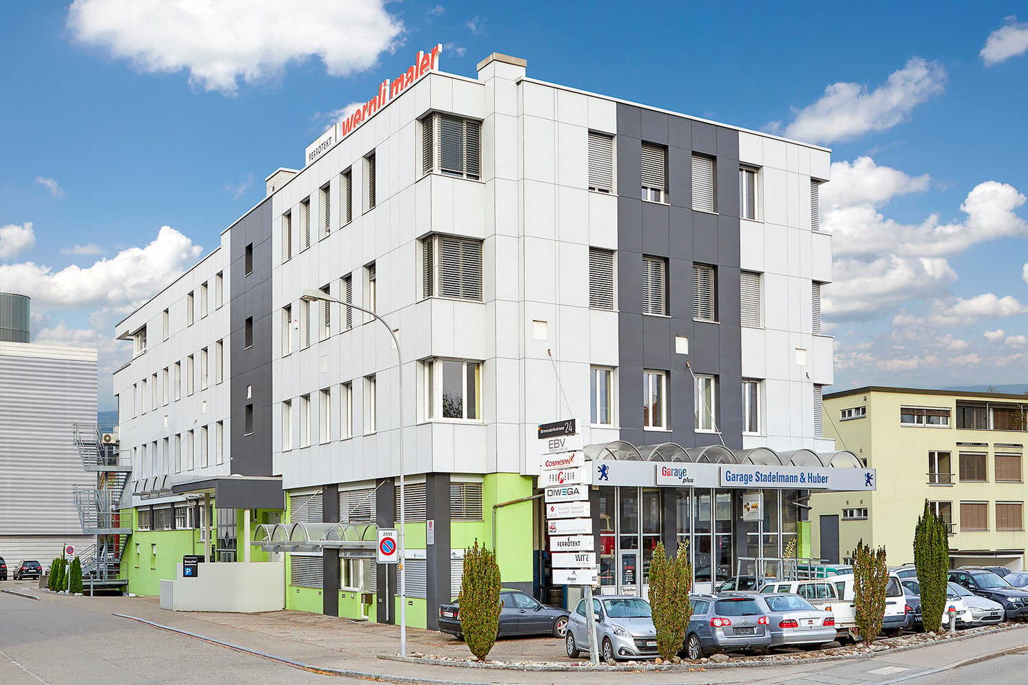 multi-story office building, white exterior, garage entrance, green accents, many windows, parked cars