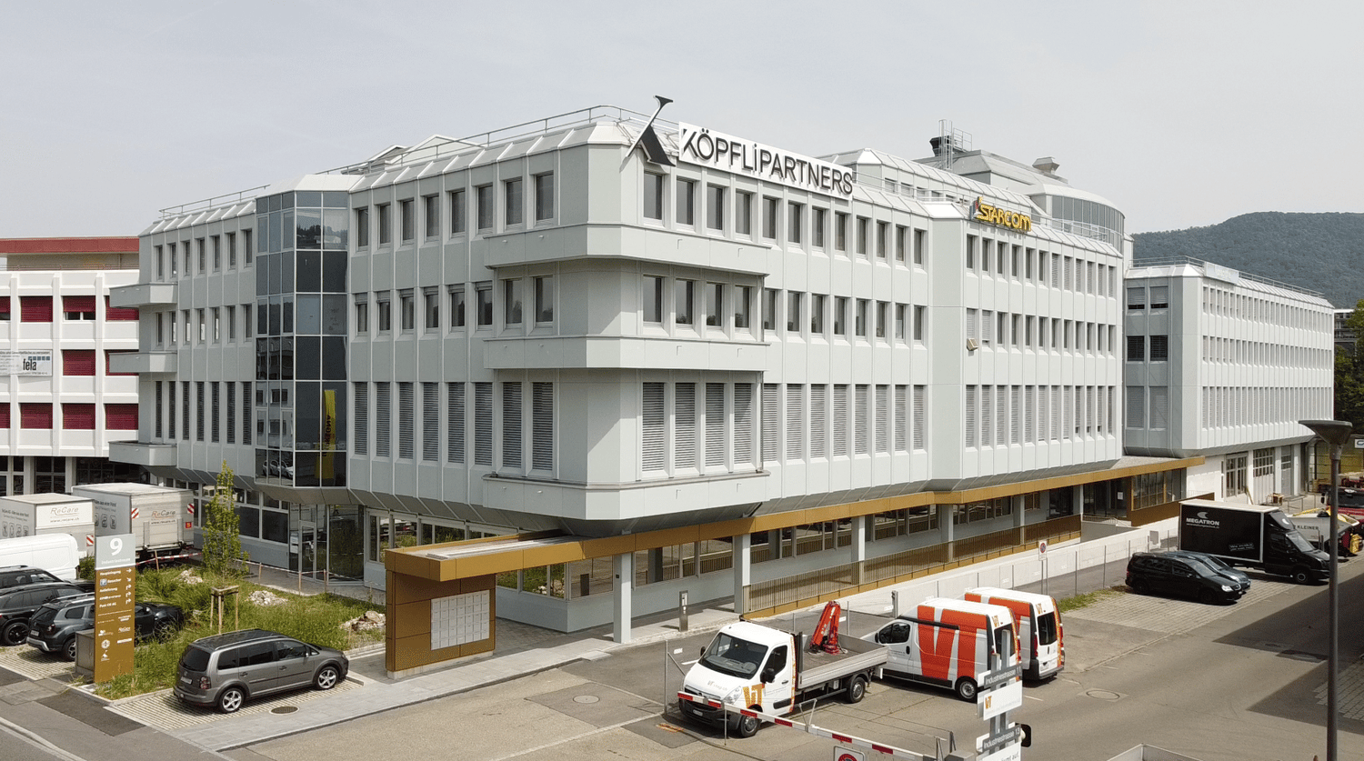 white exterior, multiple windows, 'Koepfli Partners' sign, cars parked in front, adjacent buildings, mountains in the background