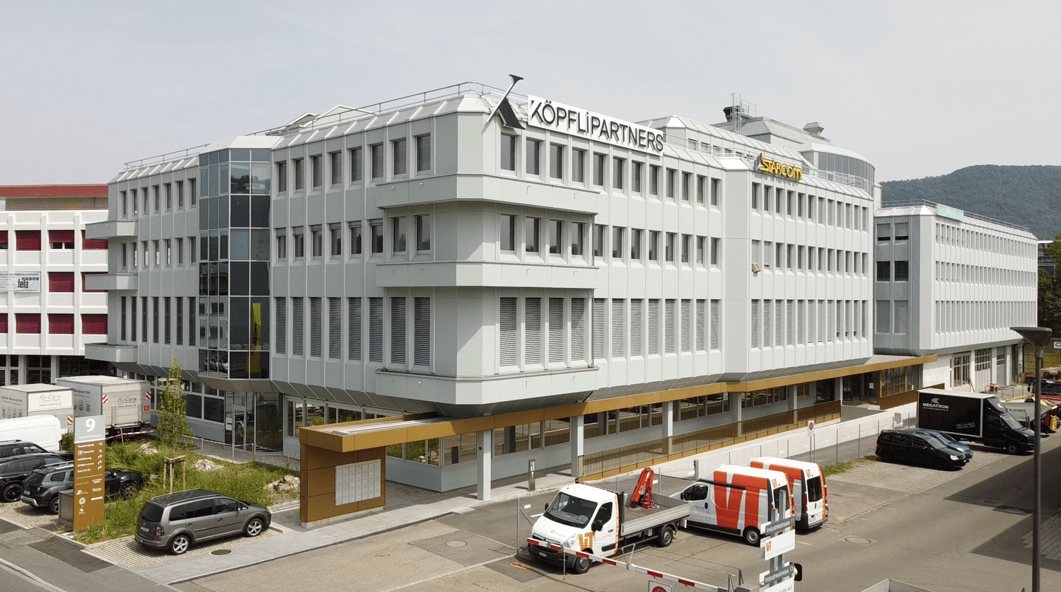 multi-story office building, white exterior, glass windows, red sign reading 'KOPFLI PARTNERS', multiple parked vehicles in the front