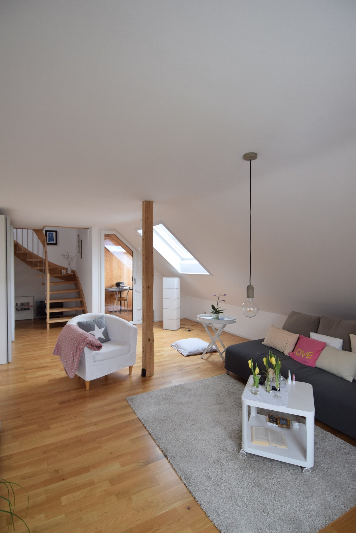 Spacious attic living room with hardwood floors, white walls, large windows, a gray sofa, a white armchair, a small side table, and a pendant light fixture. The room also features a staircase leading to another level.