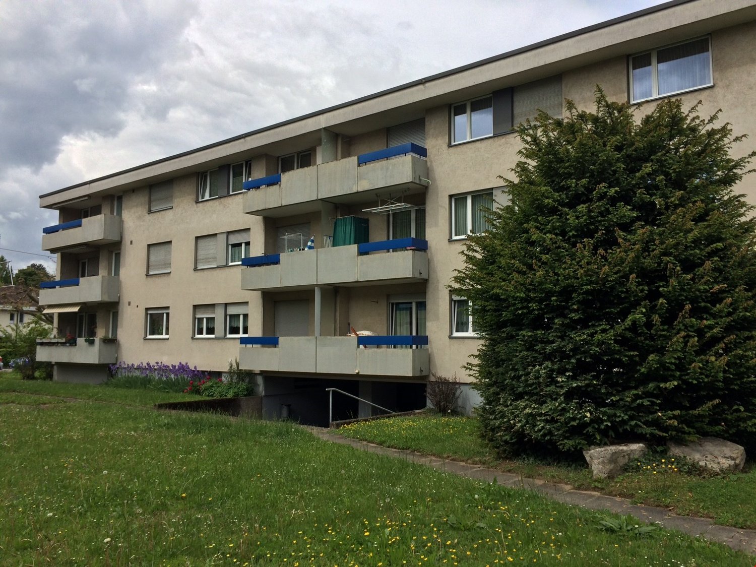 A grey concrete apartment building with multiple balconies, paved walkways, and a grass lawn.