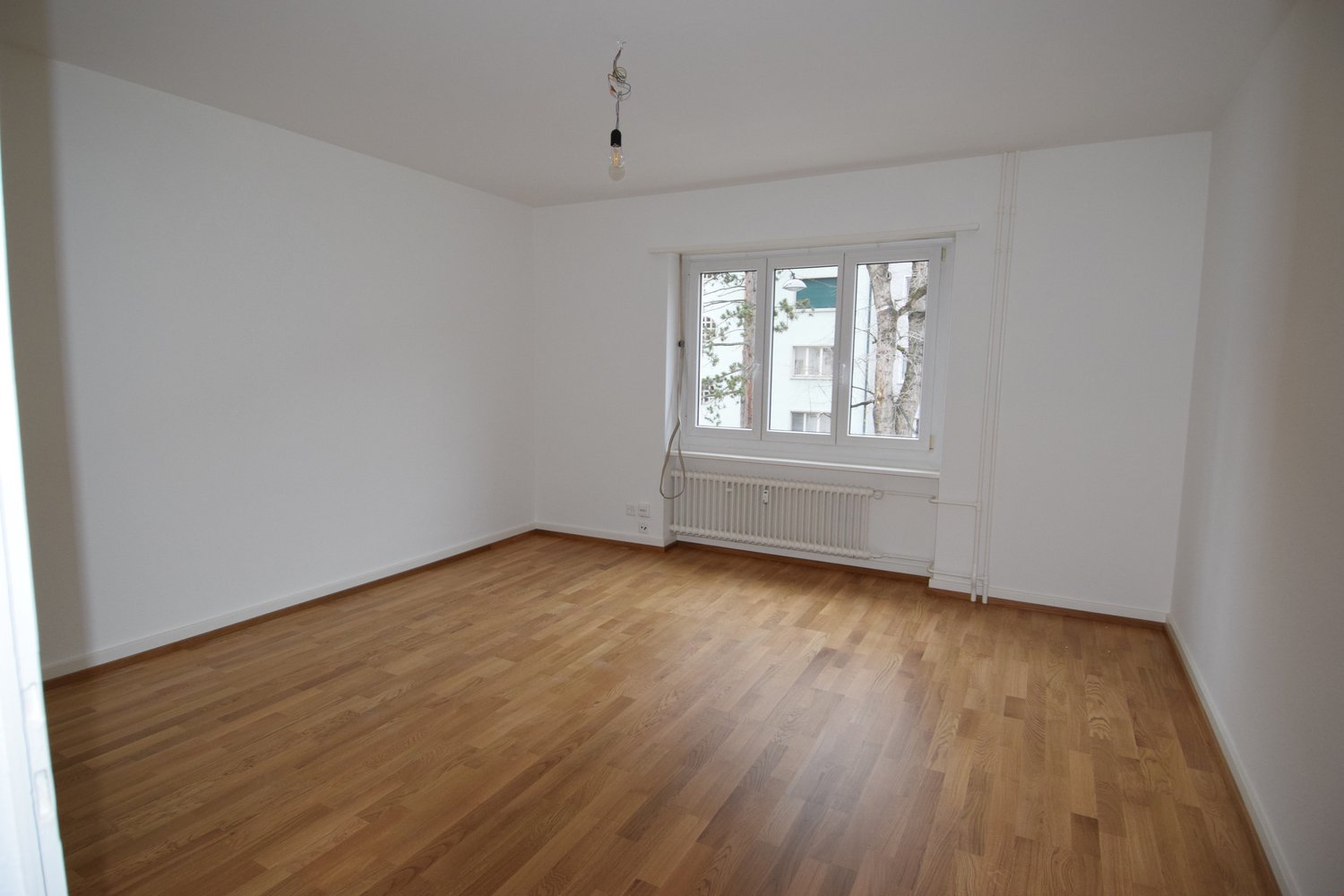 Empty room with wooden floor, white walls, large window, white radiator, single bulb ceiling light.