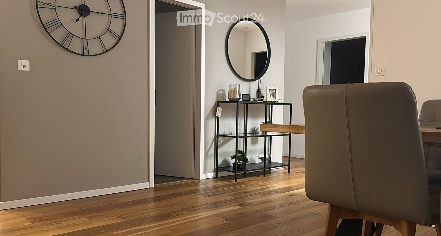 Empty room with hardwood floors, gray walls, mirror on the wall, wooden shelf with decor items, and a chair by a table.
