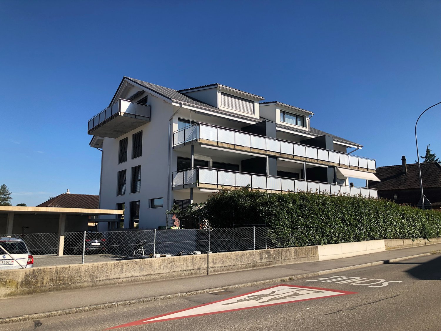 Three-story building with balconies, concrete wall with a chain-link fence, cars parked
