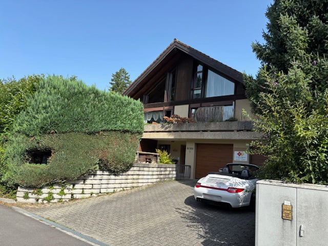 Two story modern house, brown wooden and white walls, car parked in driveway, hedges and trees around