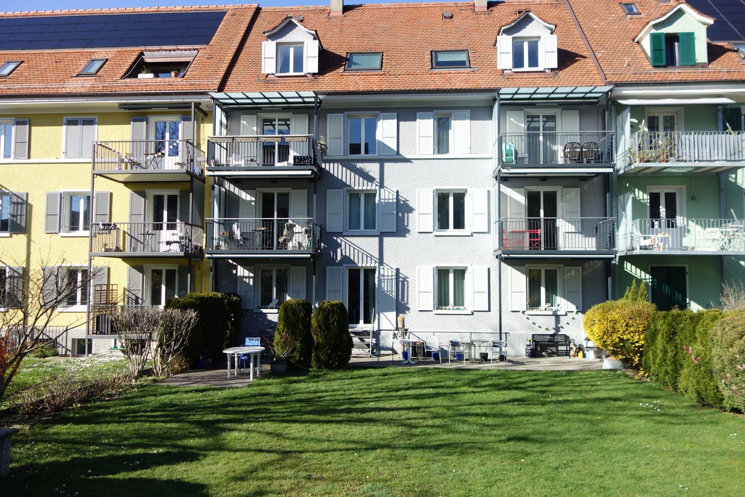 Terraced apartment building with solar panels, raised ground floor, balconies, and a garden